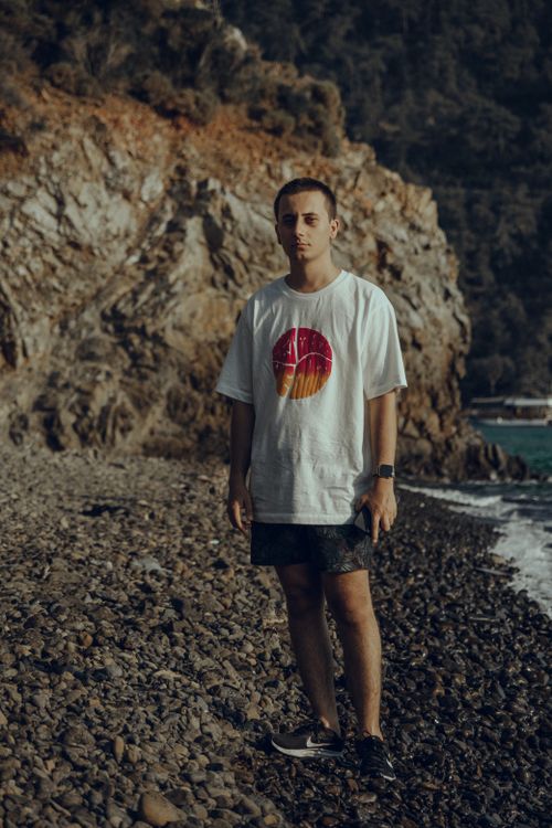 Free A young man standing on a rocky shoreline wearing a graphic t-shirt and shorts, exuding an adventurous vibe. Stock Photo