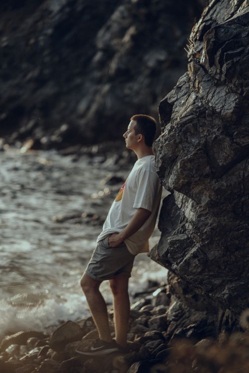 Free Young man leaning against rocks by the sea, enjoying a tranquil sunset view. Stock Photo