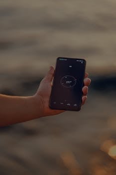 A hand holding a smartphone displaying a compass app by the water.