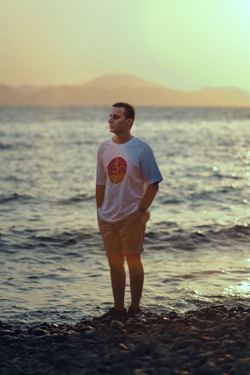 Free A man in a graphic t-shirt stands on a rocky beach, gazing at the sea during sunset. Stock Photo
