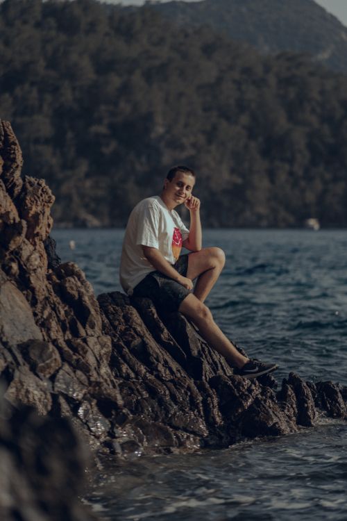 Free A man in a white t-shirt sits on rocks by the sea, exuding a calm and casual vibe. Stock Photo