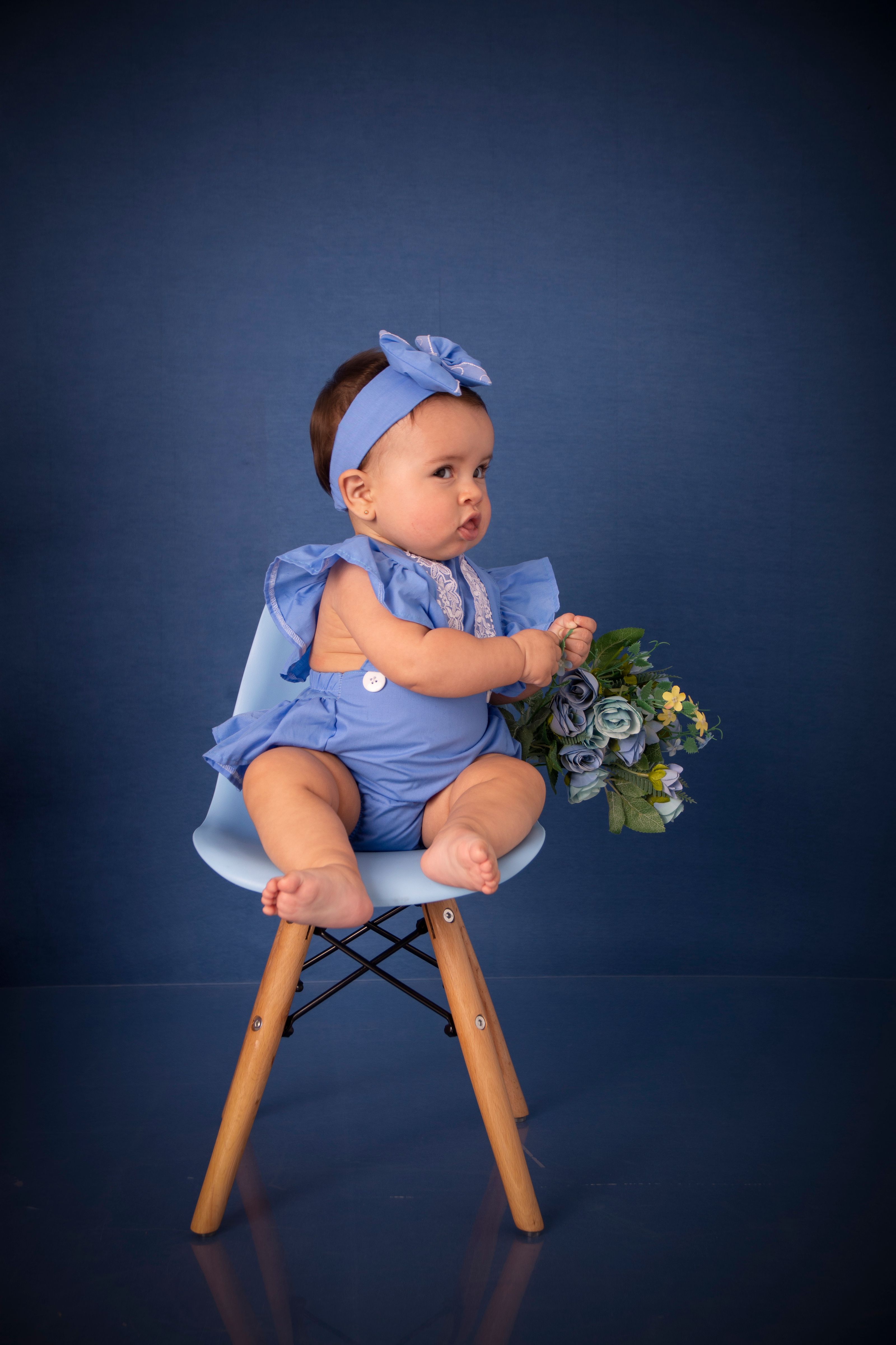 Bebê fofo sentado em uma cadeira com roupa azul e flores em estúdio.