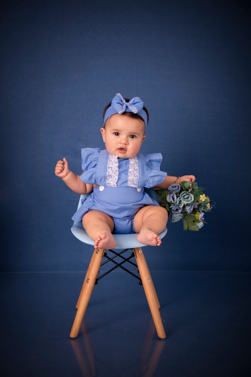 Bebê menina em traje azul sentada em cadeira decorada com flores em estúdio fotográfico