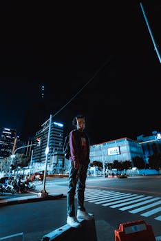 A young man standing confidently on a city street at night, with buildings and streetlights in the background.