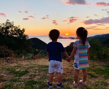 Two children holding hands, watching a beautiful sunset over the sea in Perdika, Greece.