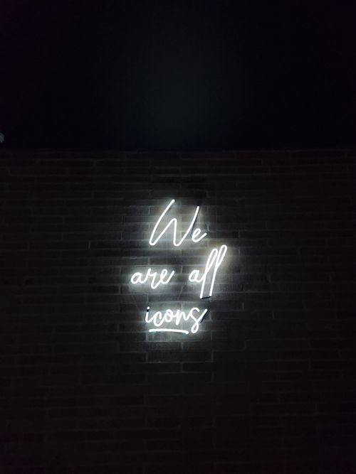 Free Bright neon light reads 'We are all icons' on a dark brick wall, photographed at night. Stock Photo