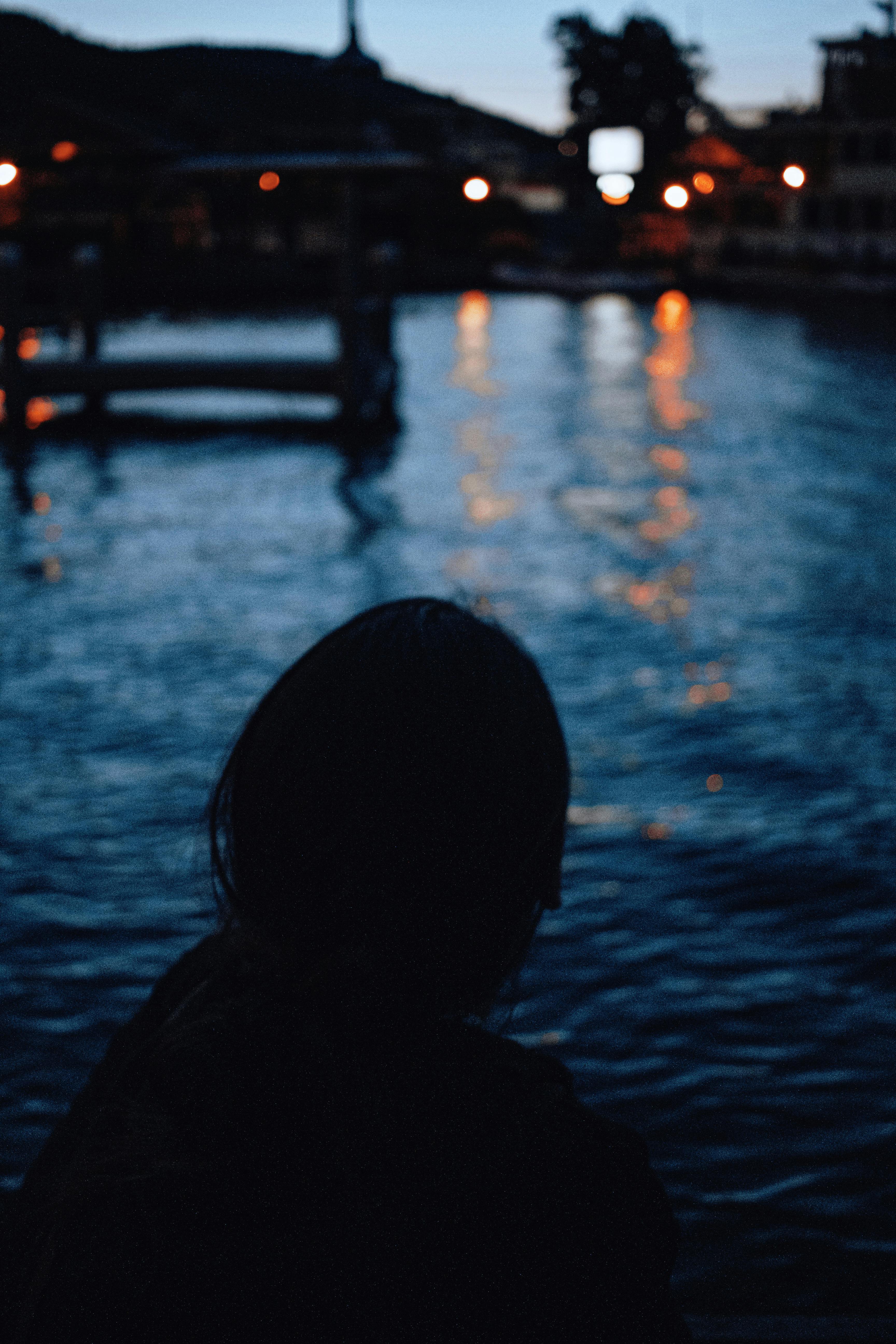 Silhouette of a person overlooking a calm marina in the evening with reflections of lights on the water.