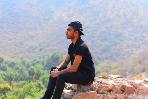 Free A young man in casual attire sits on rocks, gazing at a scenic mountain landscape. Stock Photo