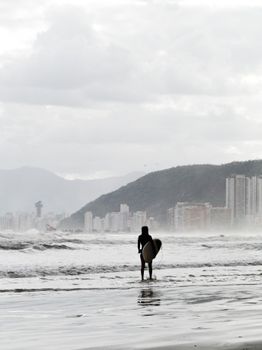A surfer stands on a beach in Santos, Brazil, ready to ride the waves with the city skyline in the background.