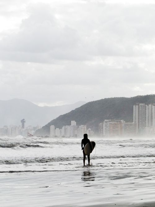 Free A surfer stands on a beach in Santos, Brazil, ready to ride the waves with the city skyline in the background. Stock Photo