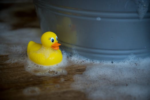 A yellow rubber duck floating near soap bubbles beside a tub on a wooden surface.