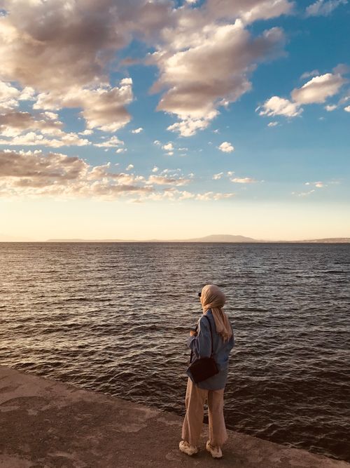 Free A woman stands by the ocean at sunset, reflecting on the vast seascape under a beautiful sky. Stock Photo