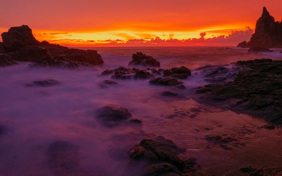 Dramatic seascape with rocks and ocean waves under a vibrant sunset glow.