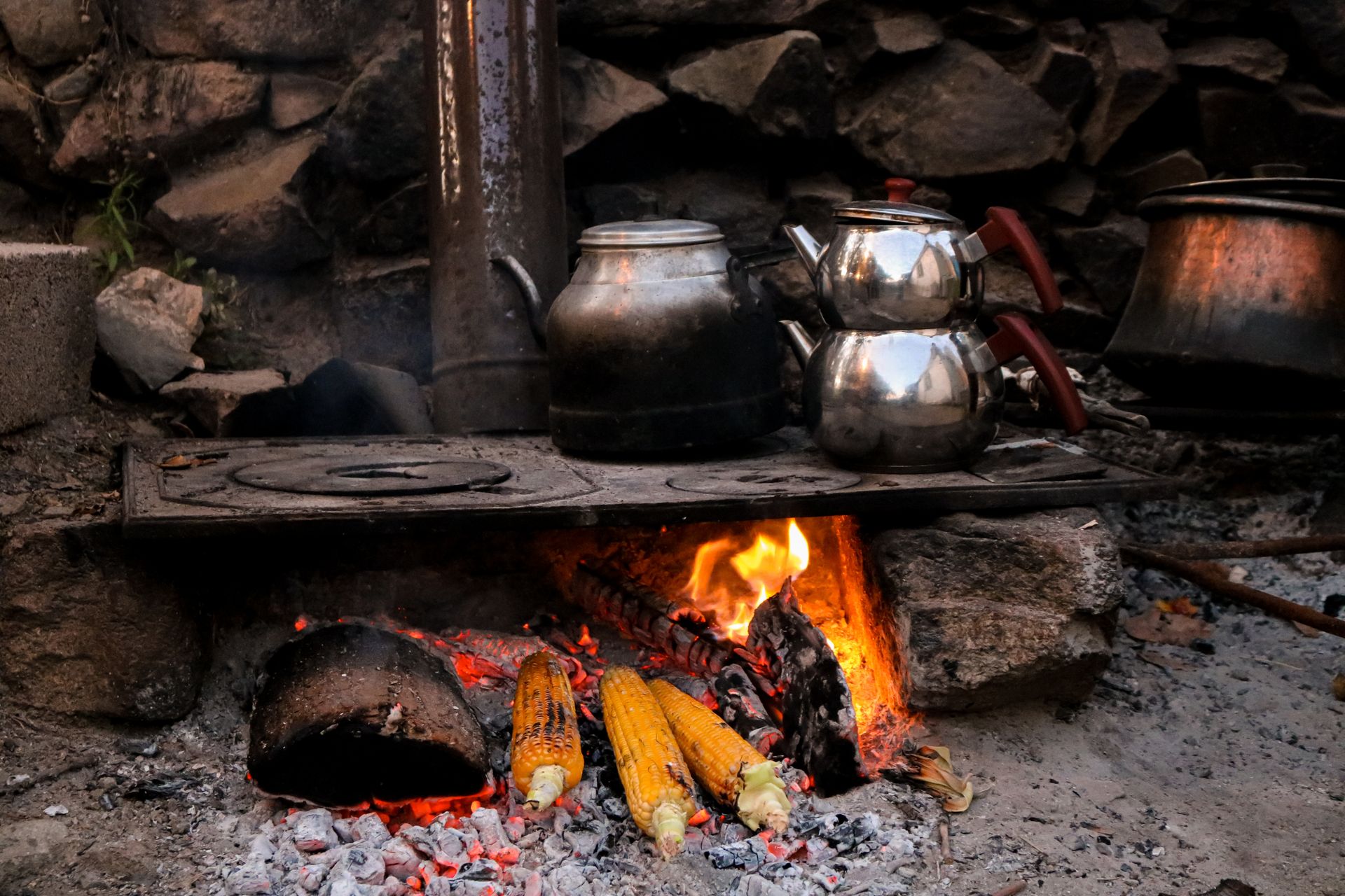 Outdoor scene of corn roasting on an open fire with metal kettles in a rustic setting.