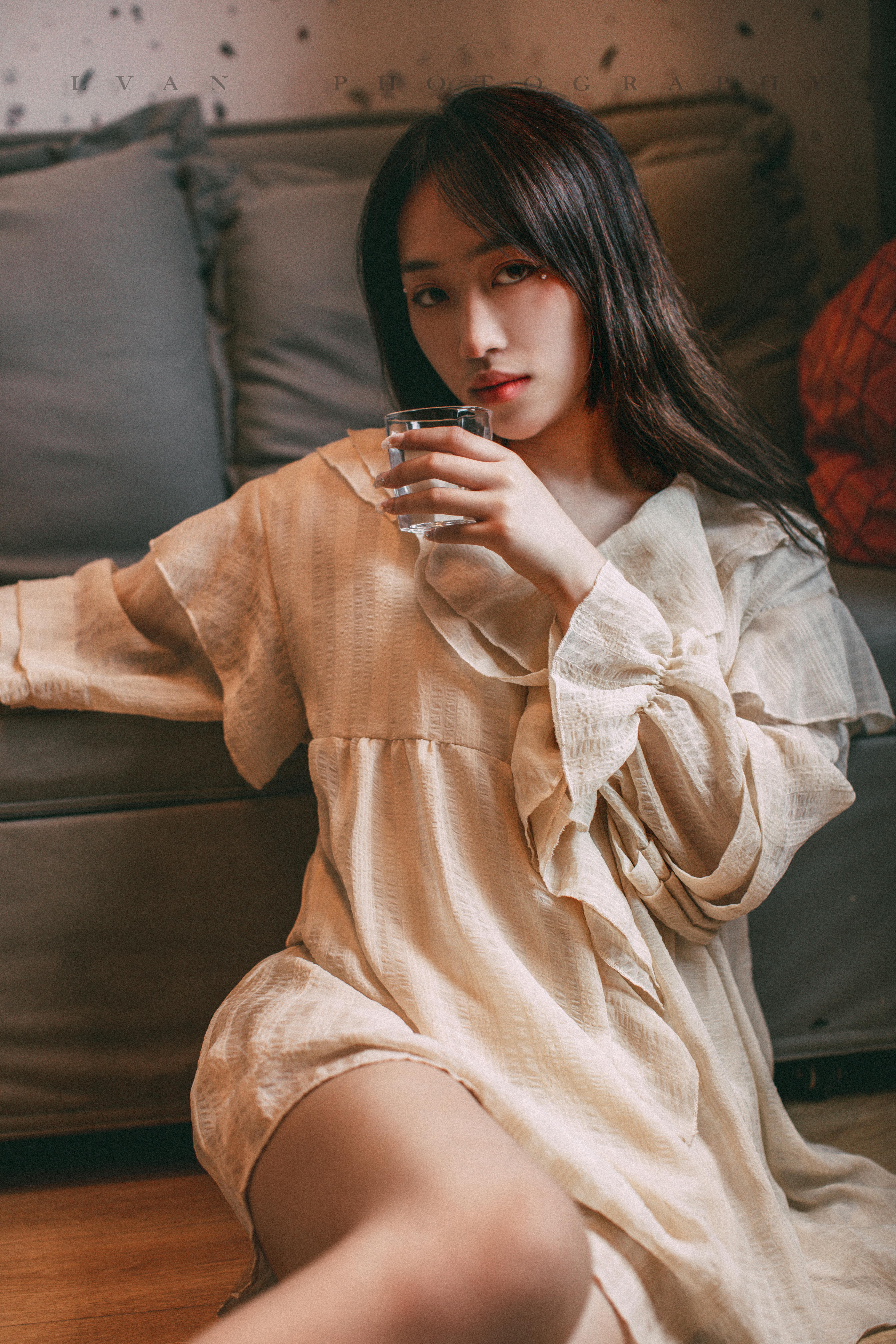 A young Asian woman in a beige dress sitting on the floor, holding a glass of water indoors.