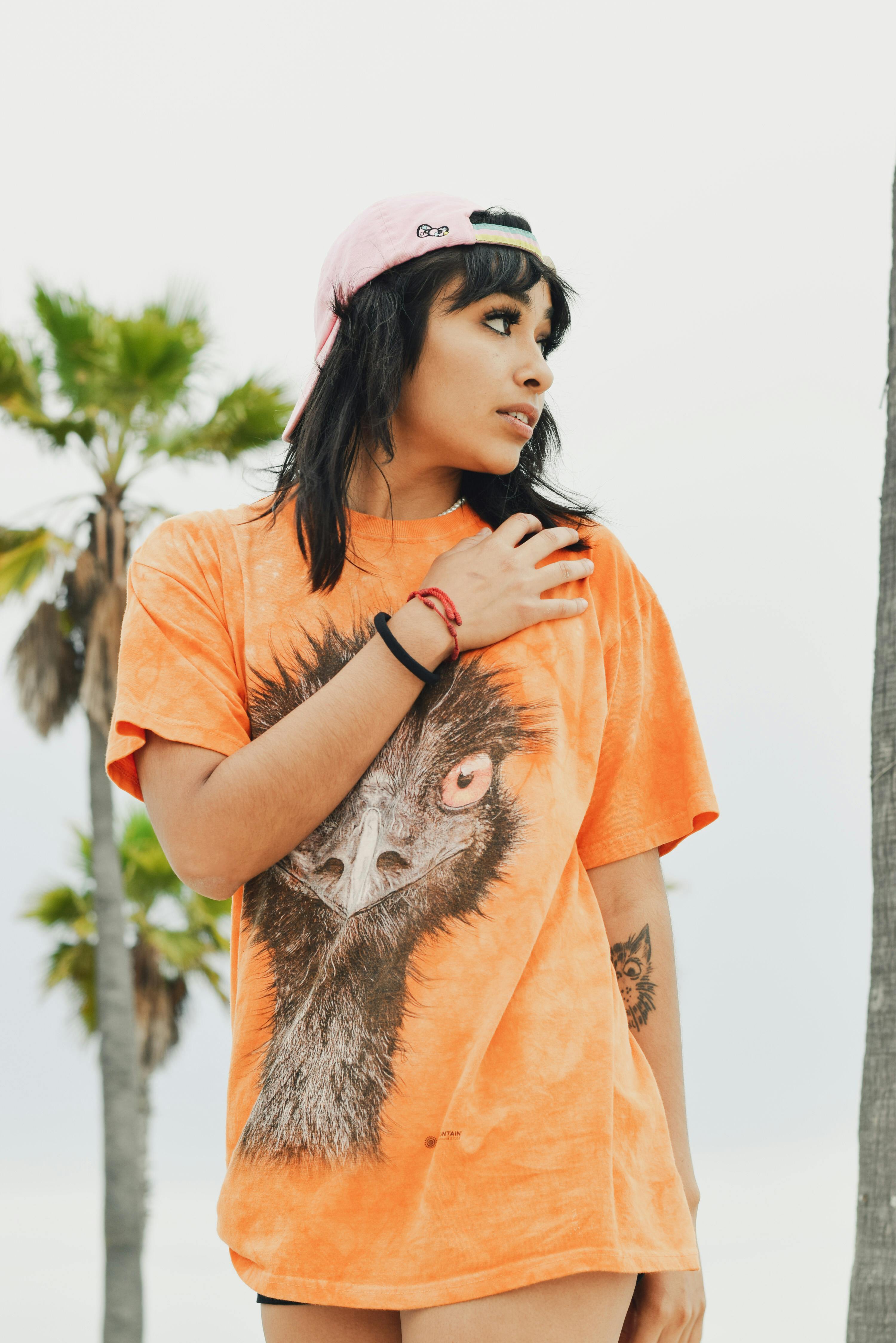 Young woman in orange graphic t-shirt and cap, outdoors in Long Beach, CA.