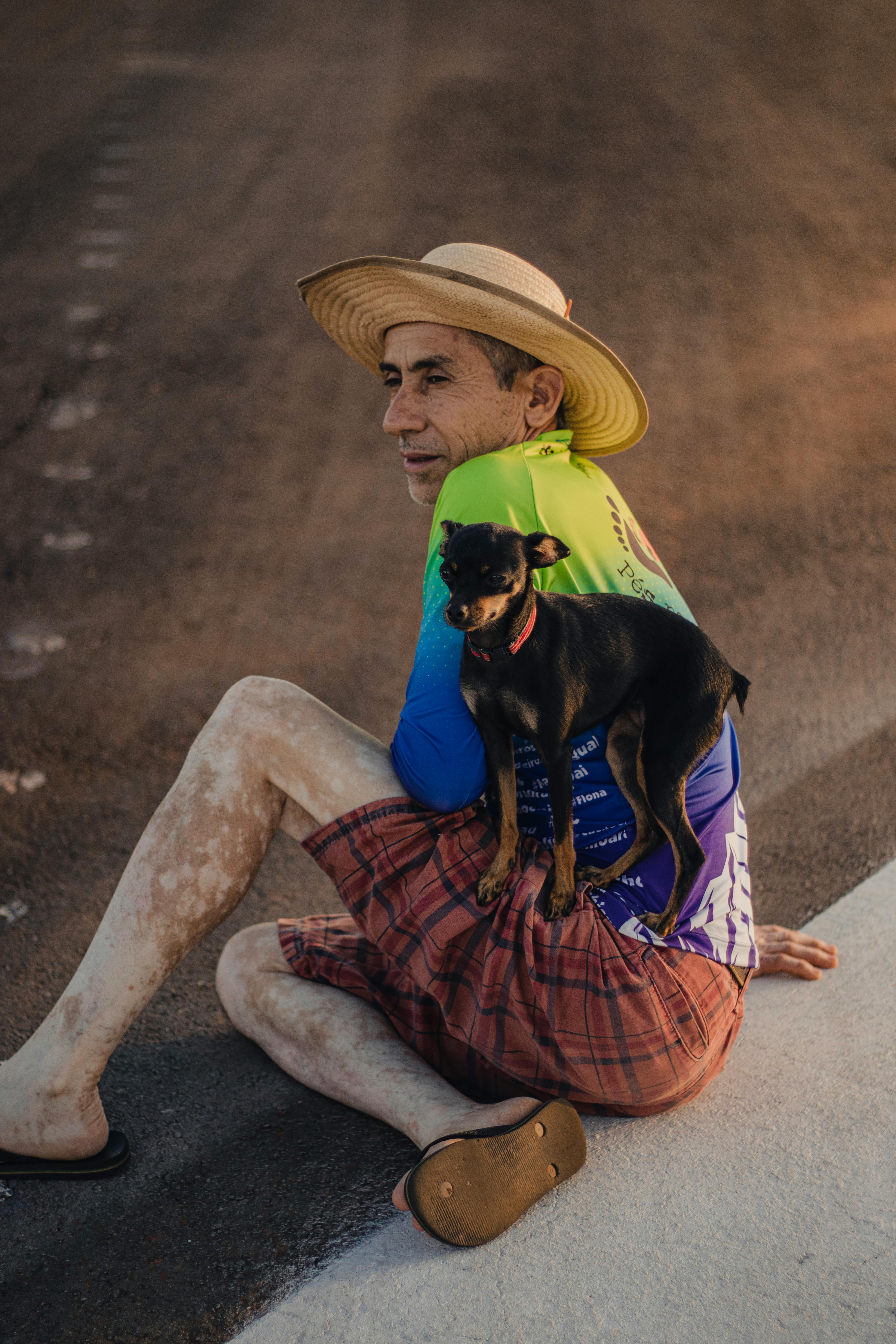 A man in a straw hat sits on a rural Brazilian road with a small black dog.