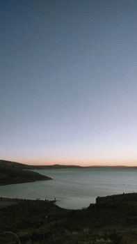 Capture the tranquillity of dusk over an expansive ocean with distant hills silhouetted against a twilight sky.