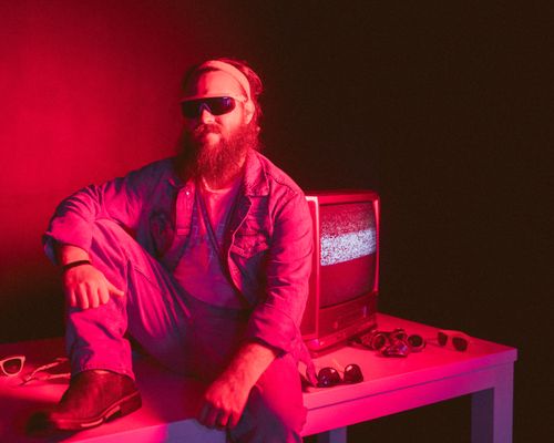 Free A bearded man in a vintage setting sits beside an old TV under pink neon lights. Stock Photo