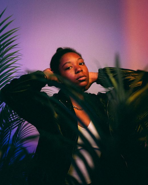 A fashionable woman poses confidently amidst vibrant indoor plants, illuminated by colorful light.