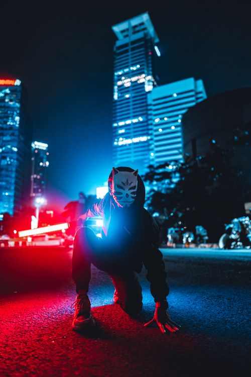 Free A masked person crouching in a vibrant cityscape at night, illuminated by blue and red lights. Stock Photo