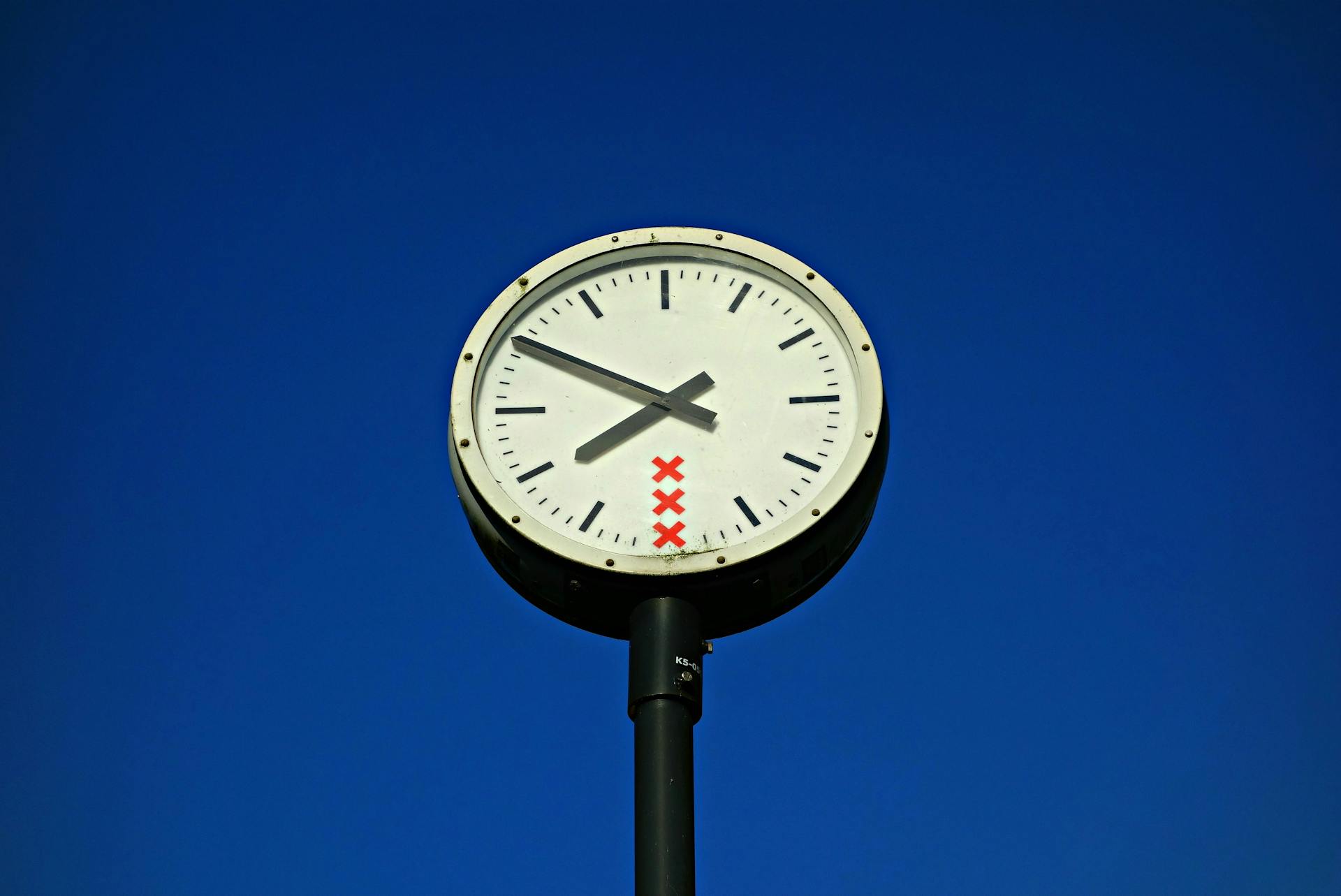 A public clock with red X's on its face stands against a clear blue sky.