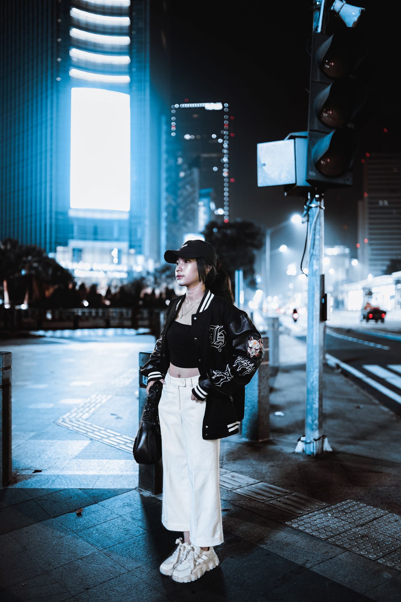 Woman in a black varsity jacket, white wide-leg pants and chunky sneakers at night