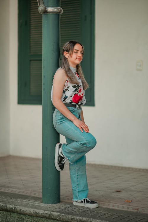 Free Woman in White and Red Floral Tank Top and Blue Denim Jeans Leaning on a Post Stock Photo