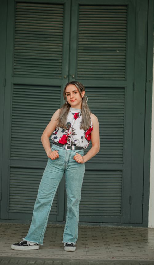 Free Woman posing in floral top and jeans stands confidently in front of a large door. Stock Photo