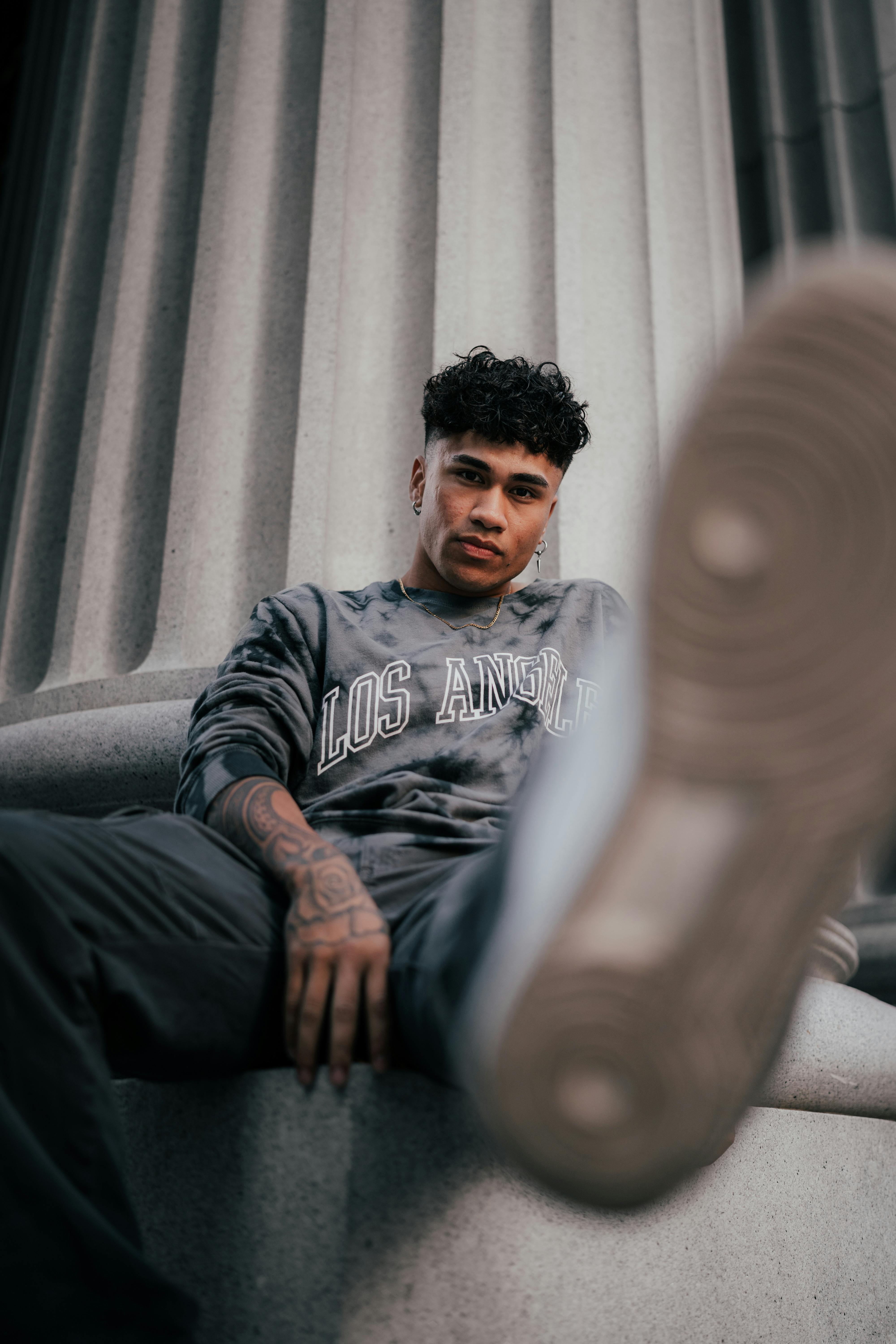 Young man in casual wear poses with a Los Angeles sweatshirt and sneakers against a column.