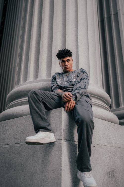 Free Stylish man in urban fashion sits by a tall column, showcasing street style. Stock Photo
