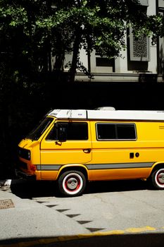 A brightly painted yellow van parked on a sunny city street under the shade of trees.