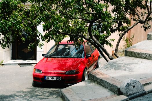 A red hatchback car parked under a lush tree in an urban environment, providing contrast and shade.