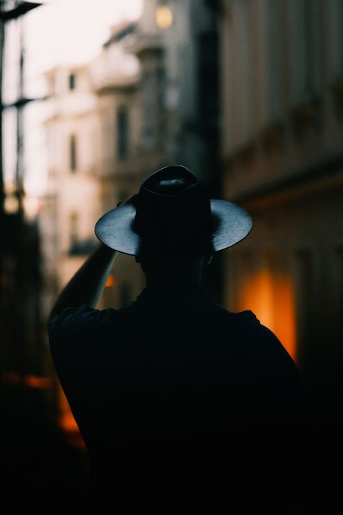 Free Backlit silhouette of a person wearing a fedora hat, capturing a mysterious urban evening scene. Stock Photo