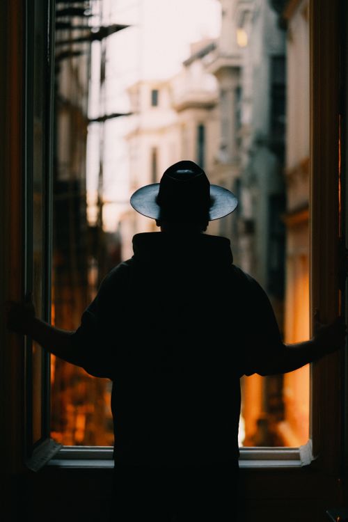 Free Silhouette of a person wearing a hat, looking out a window towards a cityscape during twilight. Stock Photo