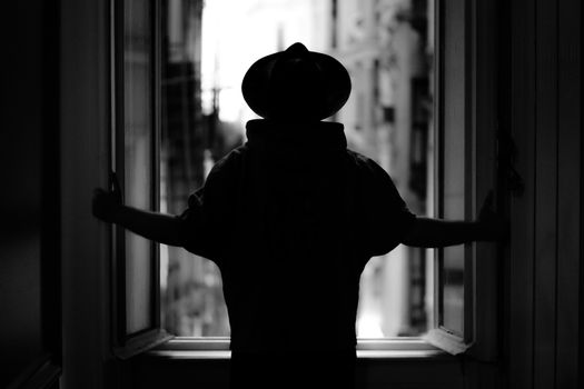 Black and white silhouette of a person in a hat at a window gazing at the cityscape.