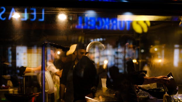 Chefs working in an ambient urban restaurant kitchen visible through a window reflection at night.