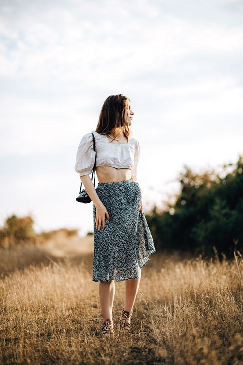 Free A stylish woman poses in a sunlit field wearing a summer blouse and skirt. Stock Photo