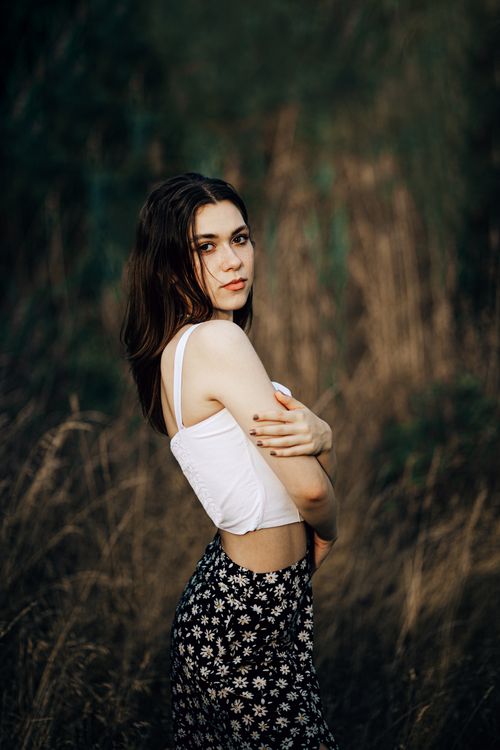 Free A woman in a white top and floral skirt poses gracefully in a grassy field during daylight. Stock Photo