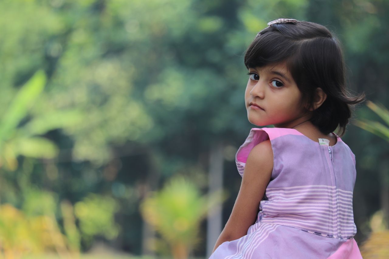 a young girl in a purple dress looks over her shoulder with a thoughtf
