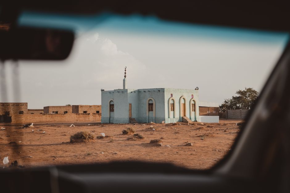 A small mosque seen through a car window in a desert landscape, under a clear sky.
