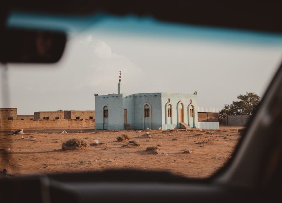 A small mosque seen through a car window in a desert landscape, under a clear sky.
