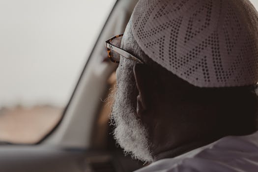 A close-up of an elderly man with glasses and kufi looking out a car window.