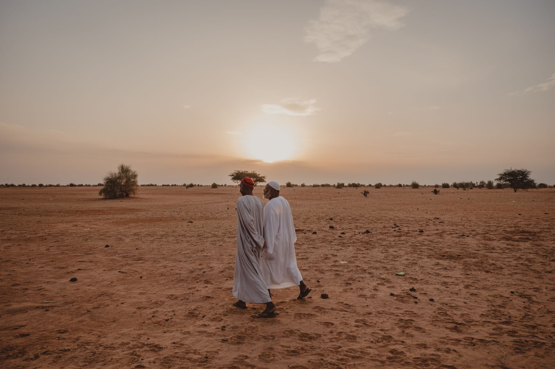 Two men in traditional attire walking in the desert at sunset.