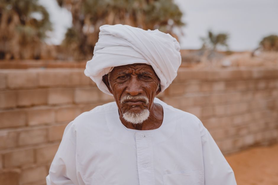Portrait of an elderly man wearing traditional clothing and a turban, standing against a desert landscape.