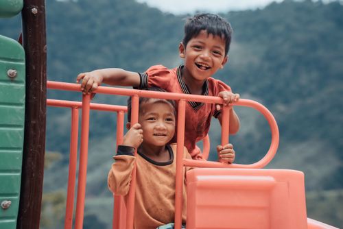 Free Two smiling children enjoying a bright day on a playground with a scenic backdrop. Stock Photo