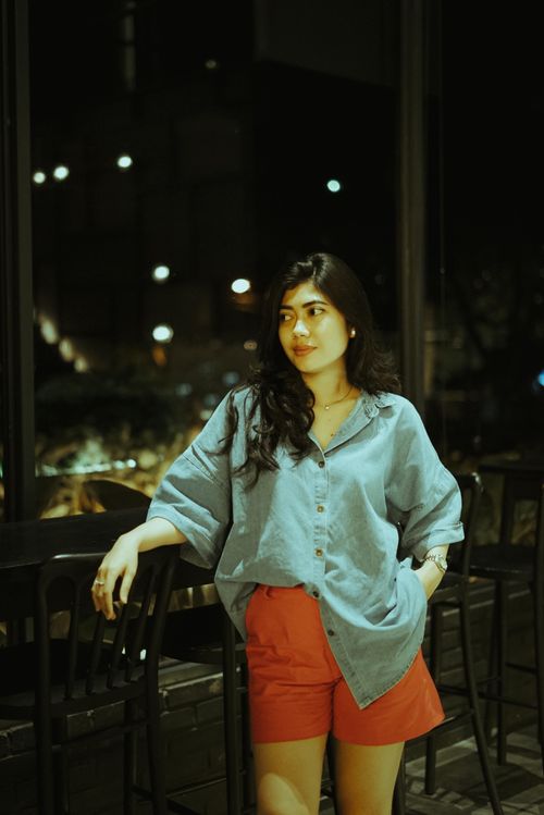 Free A young woman in casual attire, standing indoors, illuminated by warm lighting. Stock Photo
