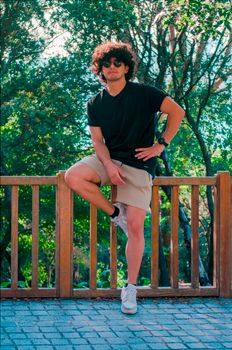 Young man with curly hair and sunglasses posing casually on a wooden fence outdoors.