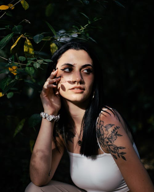 A Tattooed Woman in White Spaghetti Strap Sitting Near Green Plants while Looking Afar