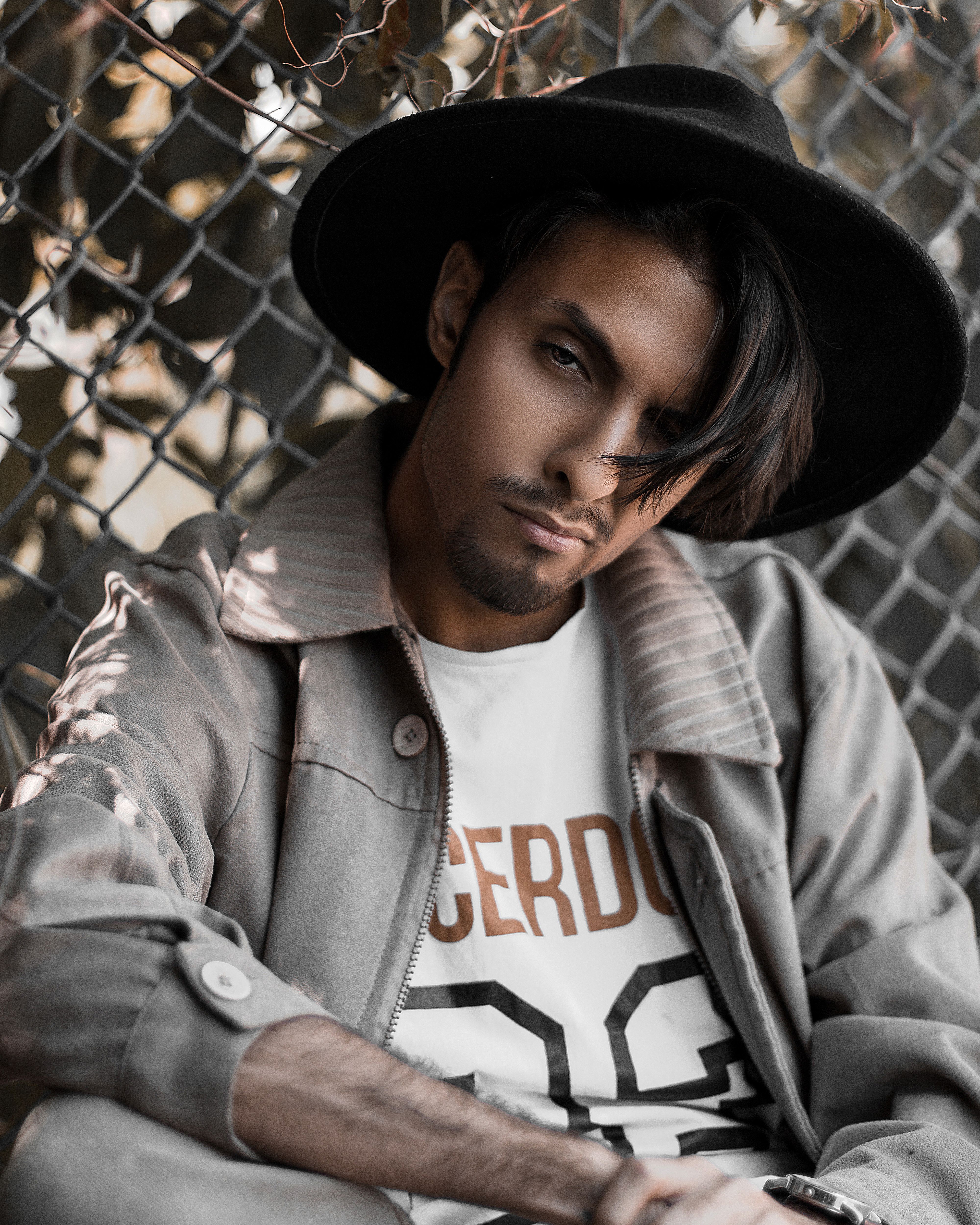 A stylish man poses outdoors in a hat and jacket against a chain-link fence.